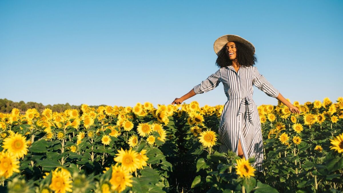 Mulher negra, de cabelo crespo e cacheado, sorri enquanto caminha no meio de um vasto campo de girassóis sob um céu azul claro. Ela usa um chapéu de palha de aba larga e um vestido longo listrado (branco e azul-marinho) com mangas e uma fenda lateral, com os braços esticados para os lados, como se estivesse aproveitando o momento. Os girassóis estão em plena floração, com as pétalas amarelas voltadas para o sol.