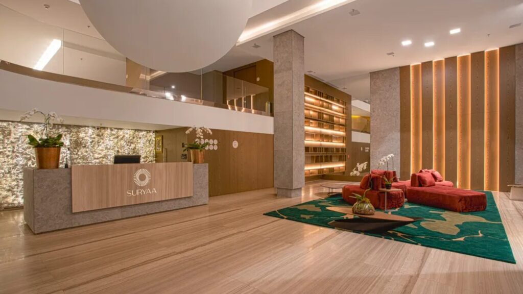 Lobby de hotel moderno e espaçoso com balcão de recepção, área de estar com sofás vermelhos e decoração em madeira e verde.
