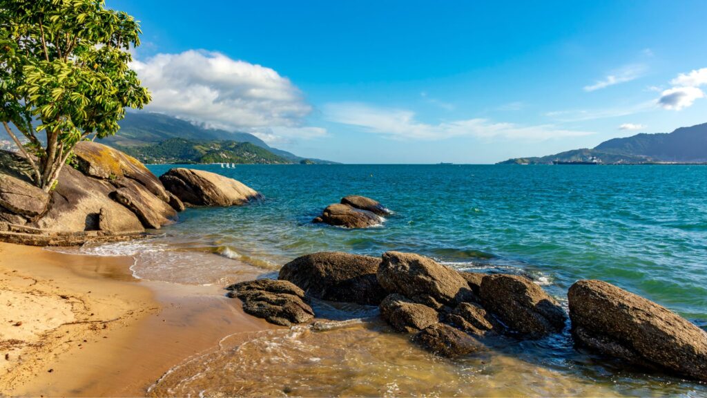 Uma vista de uma enseada rochosa em um dia ensolarado. A areia clara da praia encontra-se com as águas azuis-esverdeadas da enseada, que refletem o céu parcialmente nublado. Grandes rochas de tons terrosos estão espalhadas pela areia e avançam para a água, algumas com musgo crescendo sobre elas. Na margem esquerda, uma árvore de folhagem verde vibrante oferece sombra. Ao longe, no horizonte, uma silhueta de montanhas se eleva acima da água.