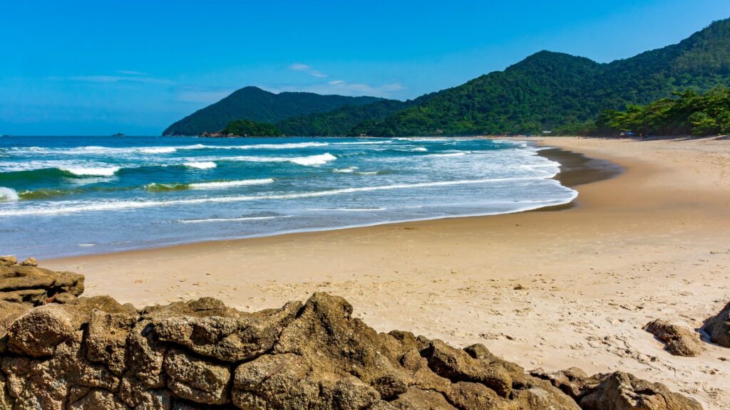 Uma vista panorâmica de uma praia tropical serena sob um céu azul claro. A areia bege-dourada se estende por toda a imagem, encontrando-se com o oceano azul-turquesa, onde ondas brancas quebram suavemente na costa. À esquerda, rochas marrons ásperas formam o primeiro plano. No fundo, montanhas verdes exuberantes cobertas por vegetação densa se elevam, dominando a paisagem.