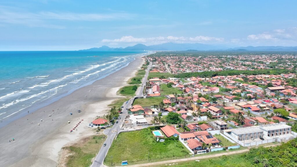 Uma vista aérea de uma extensa praia e uma cidade costeira em um dia ensolarado. A praia arenosa, de tom cinzento claro, estende-se ao longo da costa esquerda da imagem, com ondas brancas quebrando no mar azul-turquesa. Pequenas figuras de pessoas são visíveis na praia. Paralela à praia, uma estrada asfaltada serpenteia, separando a areia das casas e edifícios da cidade. As construções, predominantemente de telhados vermelhos, se espalham densamente pela paisagem à direita, estendendo-se até o horizonte. Ao fundo, uma cadeia de montanhas pode ser vista sob um céu azul claro com algumas nuvens esparsas.