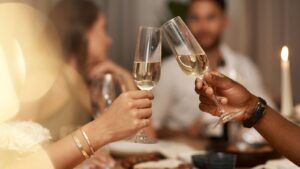 Duas pessoas fazem um brinde com taças de champanhe durante uma celebração. O foco está nas taças e nas mãos, com luzes e velas no fundo.