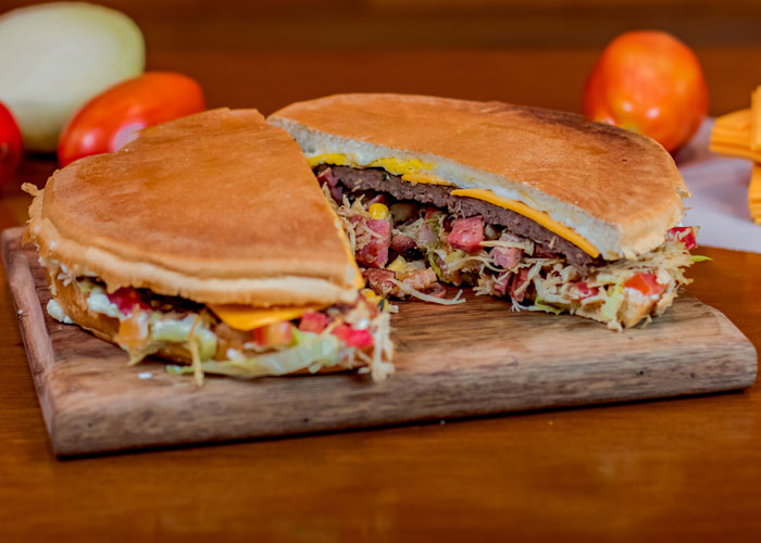 
Uma imagem exibe um XIS Gaúcho, um sanduíche típico da culinária gaúcha, cortado ao meio, revelando suas camadas internas.
