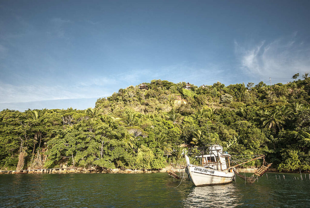 Ilha de Tinharé, com vegetação costeira e verde ao fundo. Há um barco de pesca à frente na imagem.