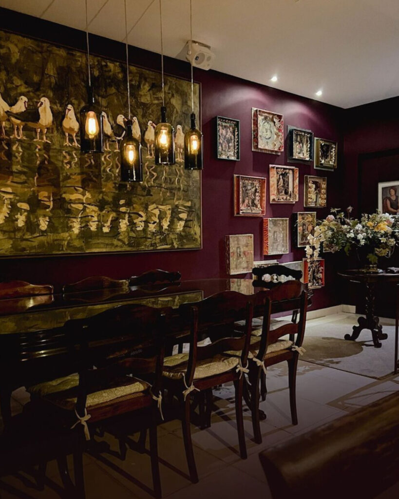 Foto do interior do Sanctuarium Atelier Gourmet. A as paredes são vermelho escuro e há muitos quadros a decorando. No canto direito da imagem, há um arranjo de flores pendentes, brancas e amarelas.