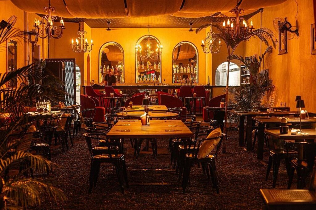 Imagem do interior do CÖA Restaurante Bar. A iluminação é laranja, assim como as almofadas das cadeiras. Há pequenas palmeiras decorativas por todo o espaço, e um bar mais ao fundo da foto.
