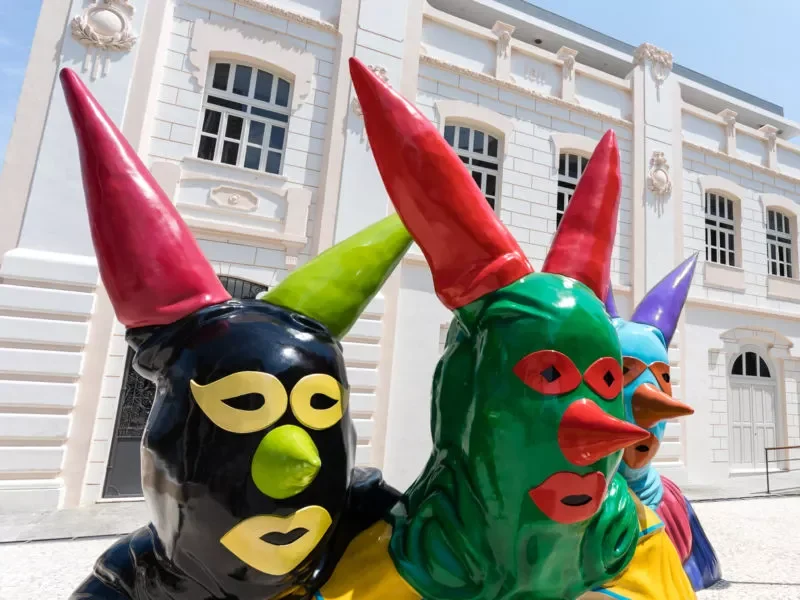 Decorações de palhaços mascarados  à frente, fachada da Casa do Carnaval atrás no plano.
