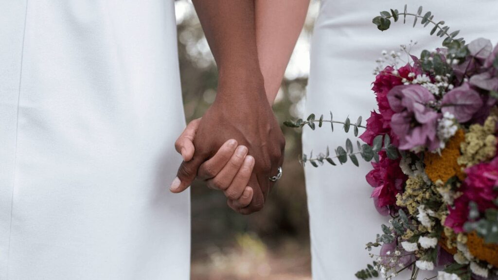 Duas noivas de mãos dadas segurando um buquê de flores.