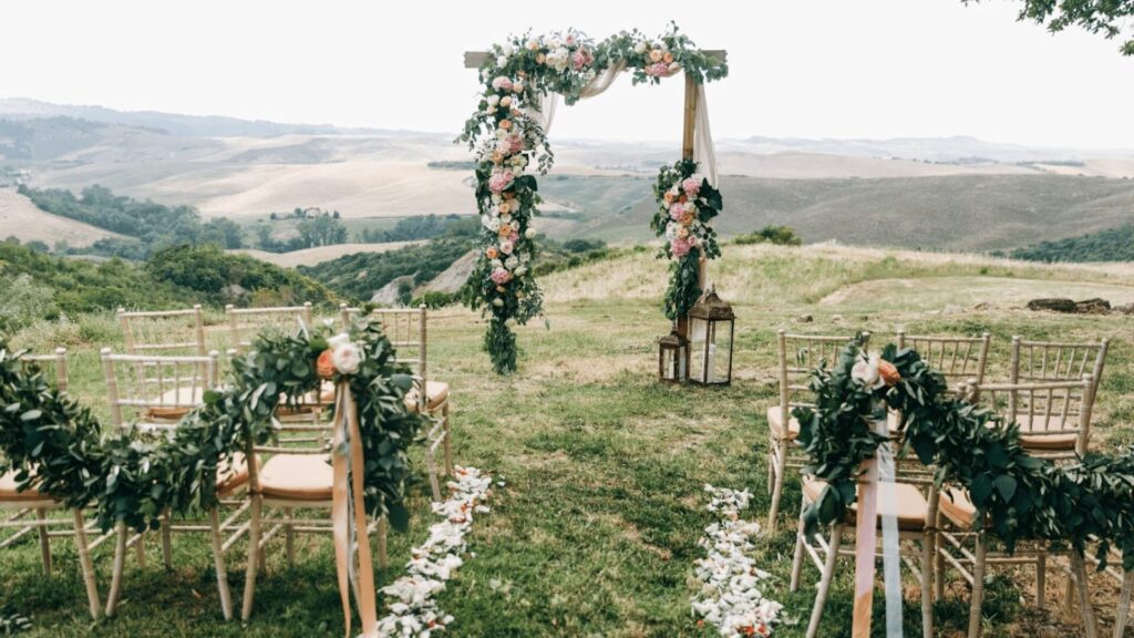 Cerimônia de casamento ao ar livre. É visto um arco retangular com tecidos fluidos e flores rosa e branca ao centro, com cadeiras bege enfileiradas.