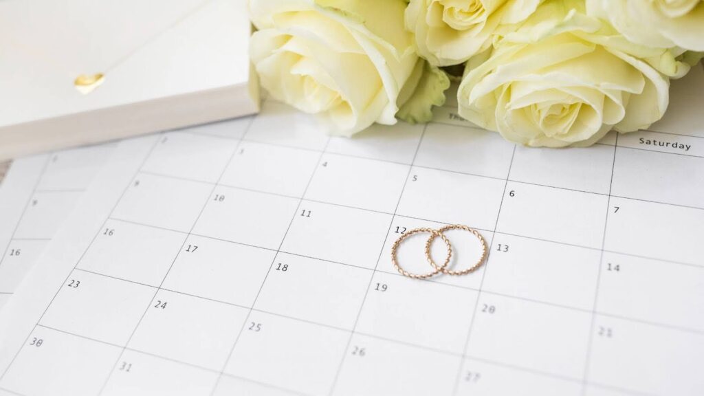 Um calendário com dois anéis dourados sobre ele, cercado por rosas amarelo pálido e um envelope branco.