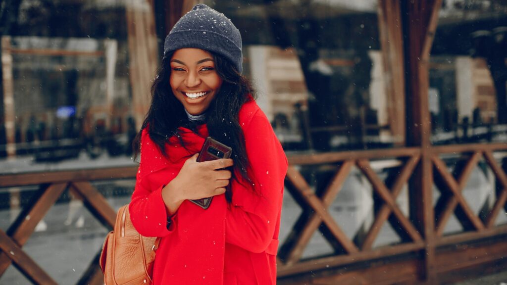 Uma mulher de casaco vermelho e chapéu sorrindo enquanto segura um celular.