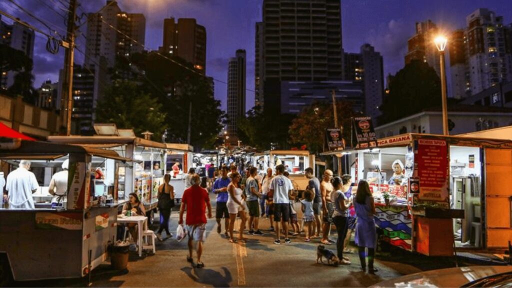 Uma movimentada rua da cidade à noite, com pedestres caminhando sob o brilho dos postes de iluminação.