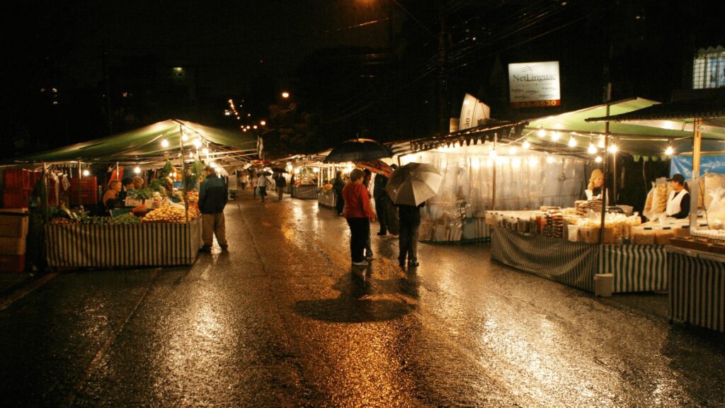 Um mercado noturno com pessoas caminhando na chuva, explorando diversas barracas e desfrutando da atmosfera.