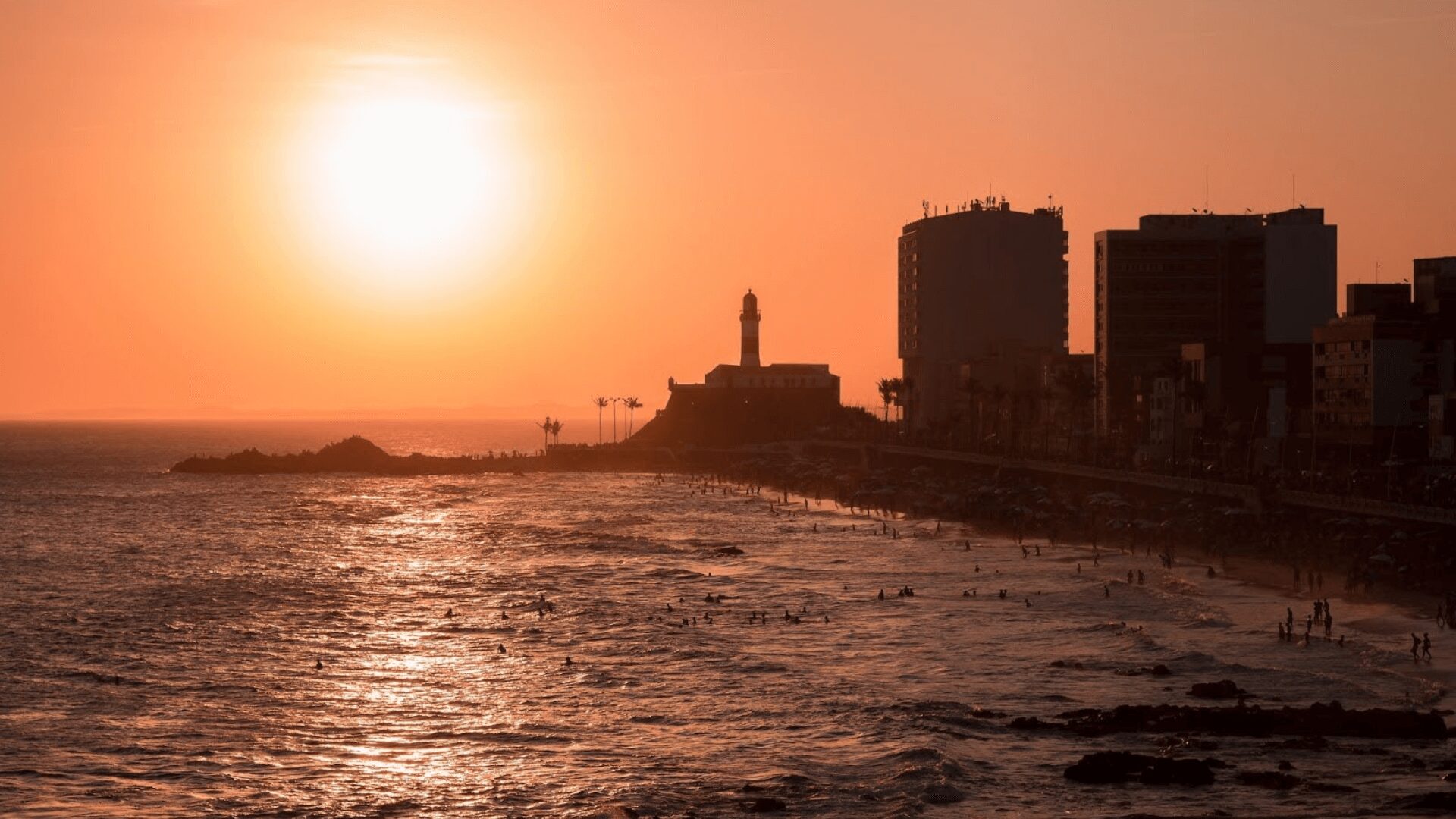 Pôr do sol sobre o oceano com edifícios ao fundo.