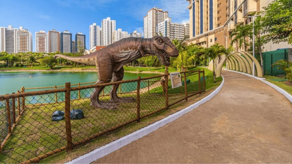 Uma estátua de dinossauro em frente a um parque com uma lagoa.