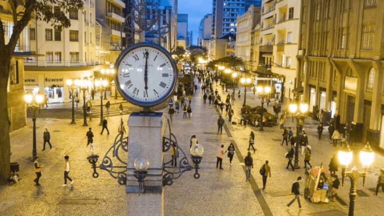 Uma movimentada rua de pedestres com uma torre do relógio em primeiro plano, rodeada por postes de luz acesos e pessoas caminhando à noite.