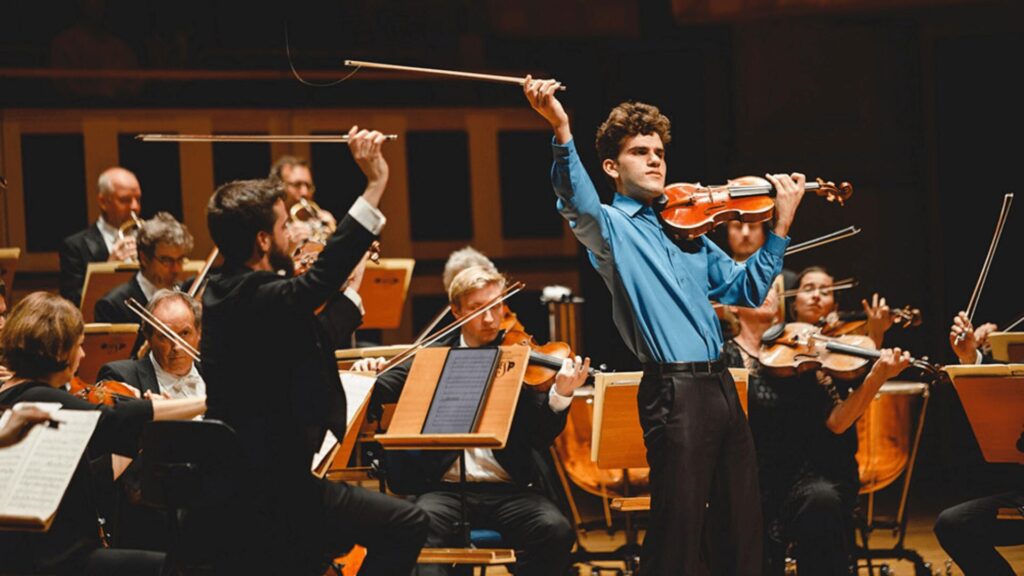 Músicos em uma orquestra tocando instrumentos de corda, vistos de trás do maestro.