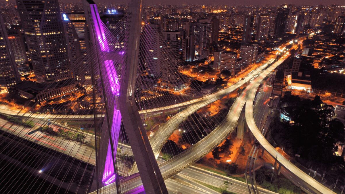 Vista aérea noturna de uma cidade com ponte roxa iluminada sobre rodovias cruzadas.