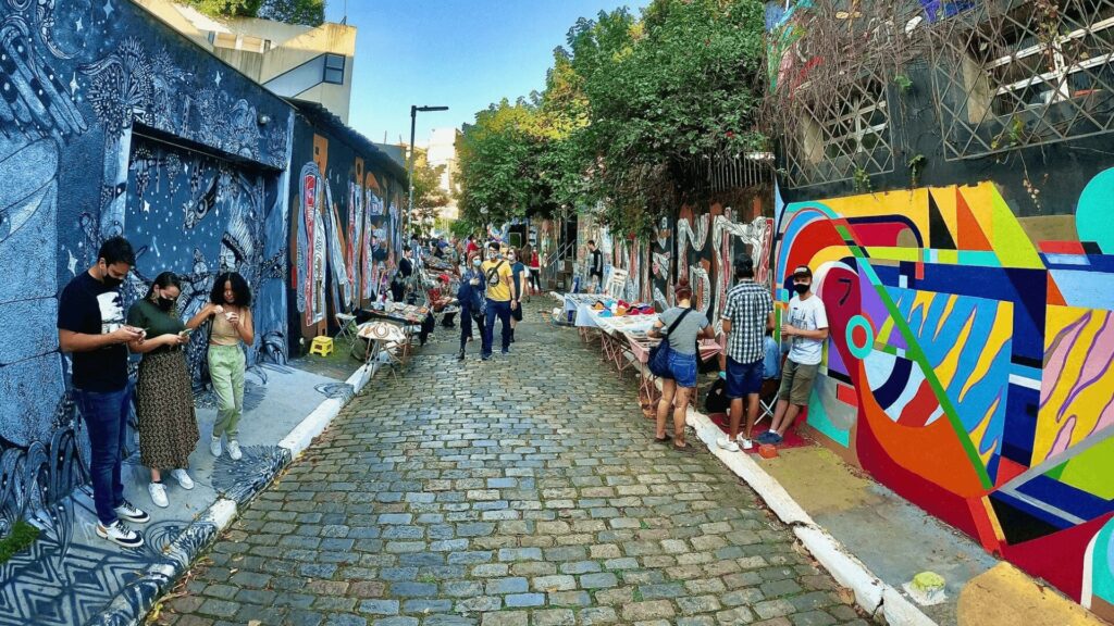 Cena movimentada de rua com pedestres caminhando ao lado de muros cobertos de grafites vibrantes.