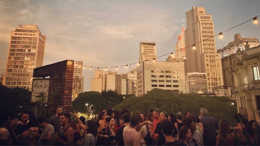 Reunião ao ar livre ao anoitecer com luzes de corda e um cenário urbano ao fundo.