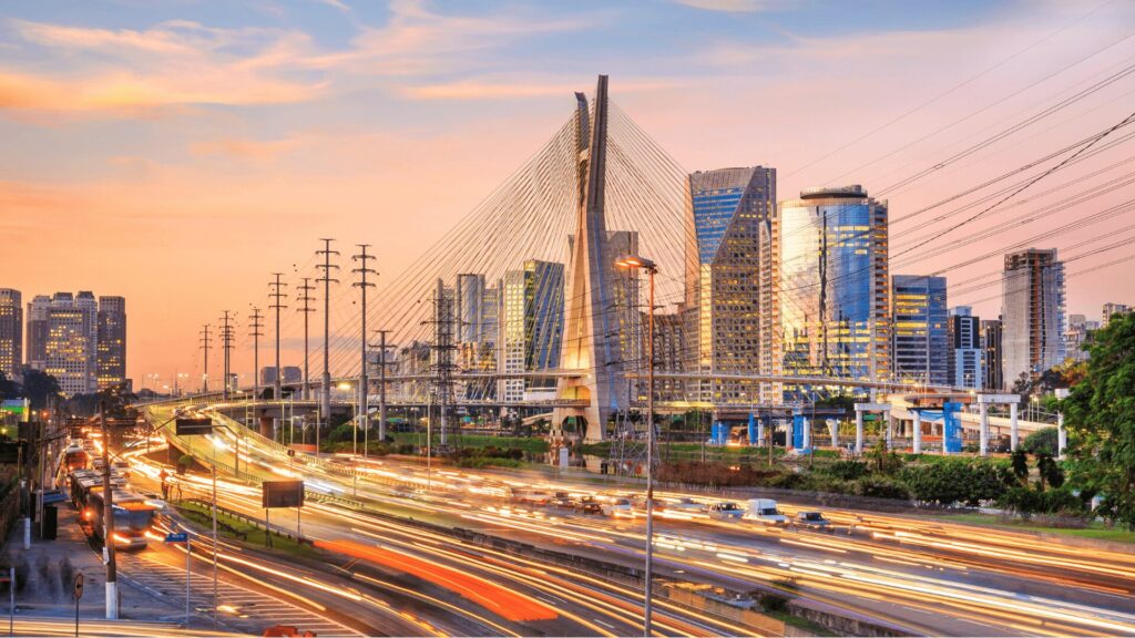 Uma rua movimentada da cidade ao pôr do sol, com tráfego e uma ponte silhuetada contra o céu vibrante.