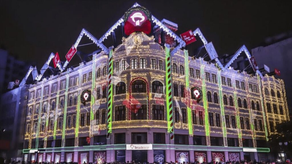 Uma multidão se reúne em frente a um edifício ricamente iluminado e decorado com enfeites festivos à noite.