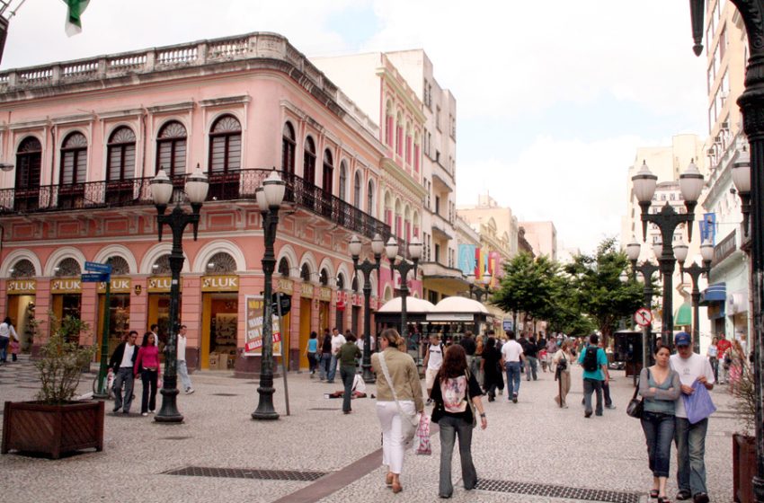 Pedestres caminhando em uma rua de paralelepípedos com edifícios históricos coloridos e postes de iluminação.