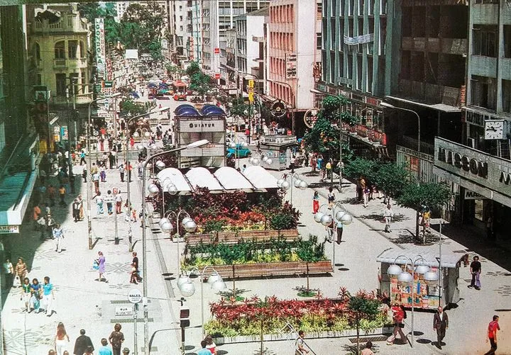 Rua urbana movimentada com pedestres, repleta de lojas e adornada com floreiras e postes de iluminação.