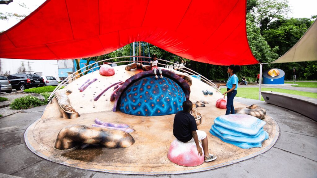 Parque infantil colorido em formato de célula, com formas abstratas e uma cobertura vermelha.