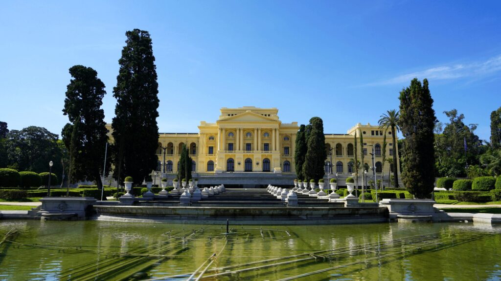 Um imponente palácio amarelo ergue-se majestosamente atrás de uma fonte e jardins meticulosamente cuidados, ladeado por árvores altaneiras que completam a paisagem grandiosa.