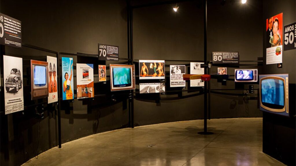 Sala de exposição com TVs e pôsteres antigos em exibição, organizados por décadas.