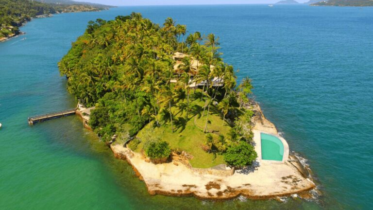 Vista de uma ilha com uma casa e uma piscina, rodeada por mar azul e palmeiras.