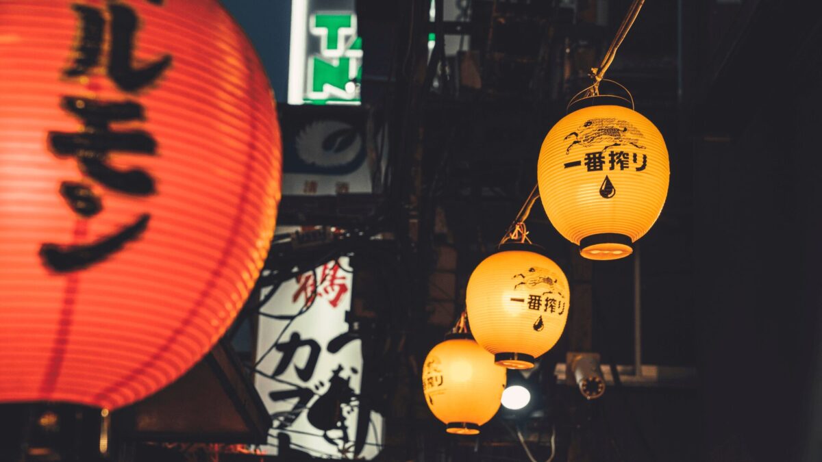 Lanternas japonesas iluminadas à noite. Uma lanterna vermelha grande com caracteres japoneses à esquerda e três lanternas amarelas menores com caracteres japoneses e o desenho de uma gota penduradas em fila à direita.