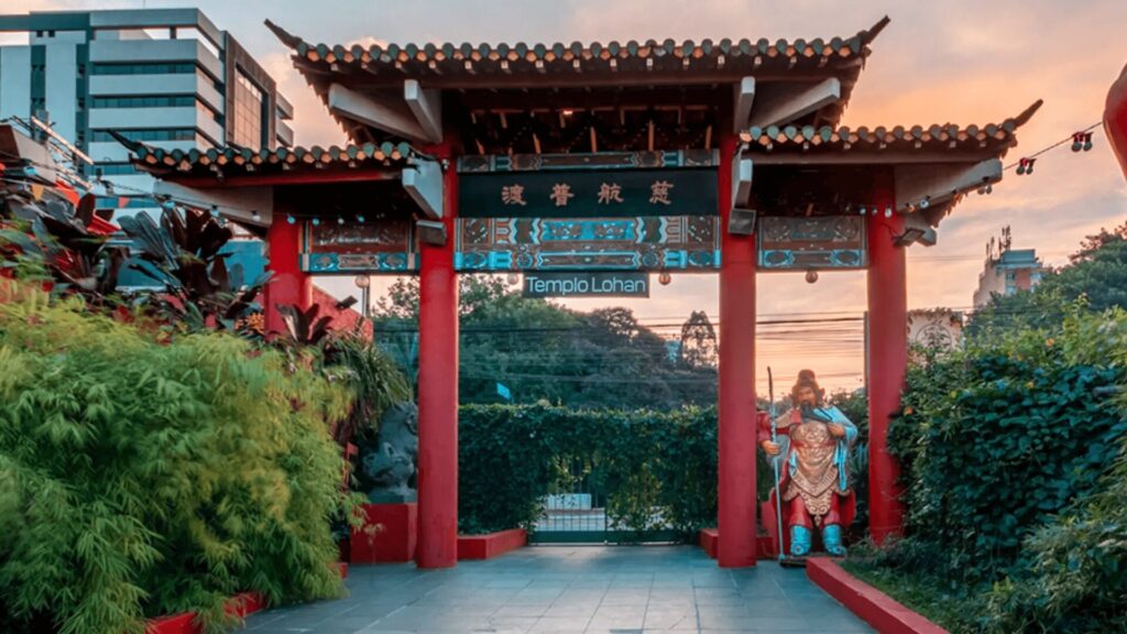 Um portal de entrada tradicional chinês com telhado de várias camadas e pilares vermelhos. Um letreiro com caracteres chineses e a inscrição "Templo Lohan" está acima do portão. Uma estátua de um guerreiro chinês com armadura e expressão imponente está ao lado do portão. Arbustos verdes e um céu alaranjado ao pôr do sol compõem o fundo.