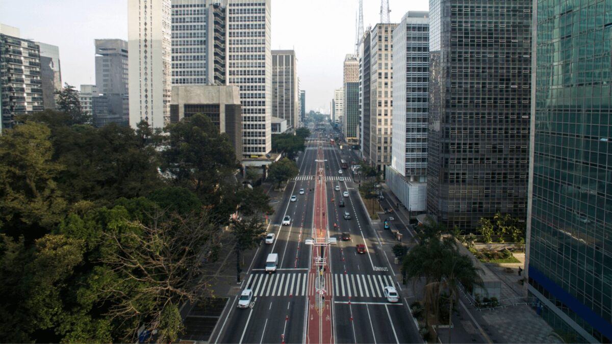 Vista aérea da Avenida Paulista, em São Paulo, com prédios altos e modernos dos dois lados. A avenida está movimentada com carros, ônibus e pedestres atravessando a faixa de pedestres. Há árvores e áreas verdes ao longo da avenida, contrastando com a paisagem urbana.