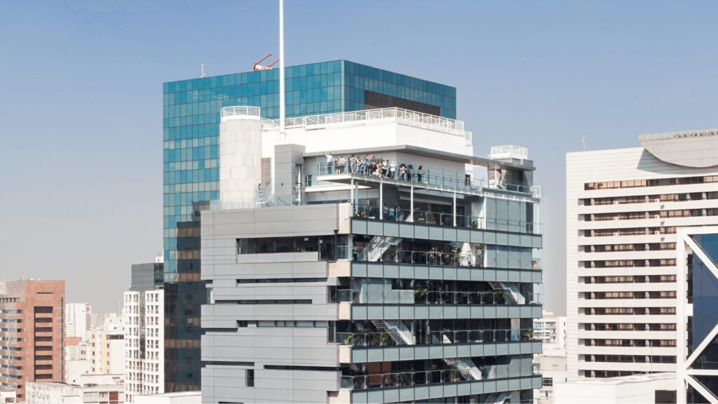 Vista da cobertura do Sesc Avenida Paulista, com a bandeira brasileira tremulando ao vento. Várias pessoas estão reunidas no terraço, apreciando a vista da cidade. O edifício moderno se destaca na paisagem urbana com sua arquitetura imponente e janelas espelhadas.