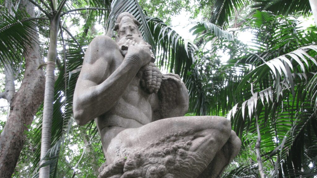 Uma estátua de pedra de um fauno, uma criatura mítica. Ele está ajoelhado, com uma barba longa e chifres. A estátua está localizada em um parque exuberante, com palmeiras e outras folhagens ao redor.