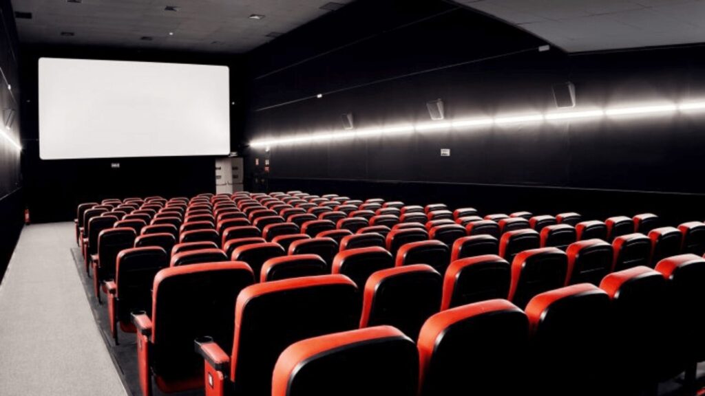 Interior de uma sala de cinema vazia com fileiras de poltronas vermelhas voltadas para uma grande tela branca em branco.