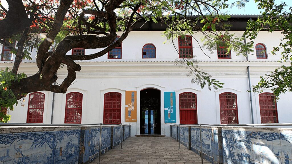 Entrada de edifício colonial branco com portas vermelhas e azulejos azuis, ladeado por vegetação.