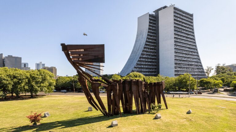 Uma escultura abstrata de metal enferrujado, lembrando uma caravela estilizada, destaca-se em um gramado bem cuidado. Ao fundo, um moderno prédio com fachada curva e janelas espelhadas contrasta com a escultura e a vegetação ao redor.