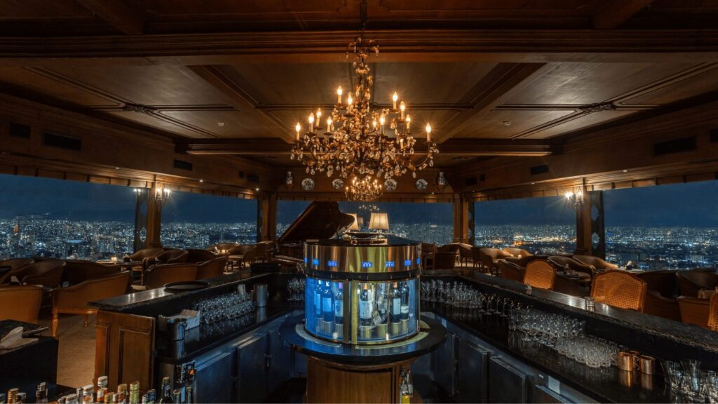 O interior luxuoso e aconchegante do bar do Terraço Itália, com um lustre ornamentado, um piano de cauda e janelas panorâmicas com vista para o horizonte noturno da cidade de São Paulo.