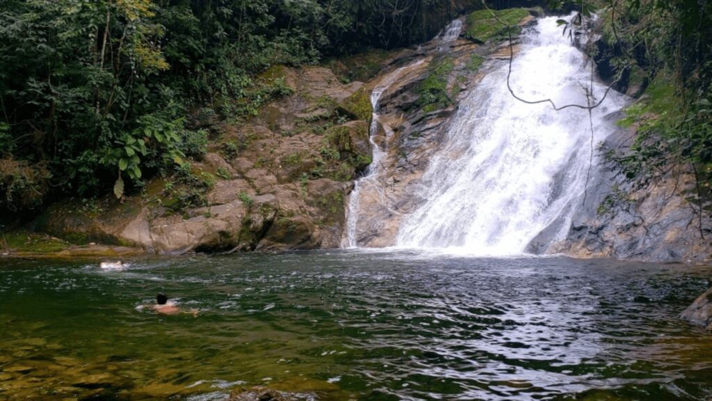 Uma cachoeira em cascata desce por um conjunto de rochas, criando várias quedas d'água menores. A vegetação exuberante e uma ponte de madeira rústica adicionam charme ao cenário.