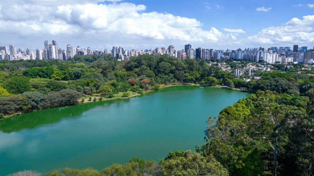 Uma vista panorâmica do Parque Ibirapuera em São Paulo, mostrando um lago sereno cercado por árvores exuberantes e prédios da cidade ao fundo.