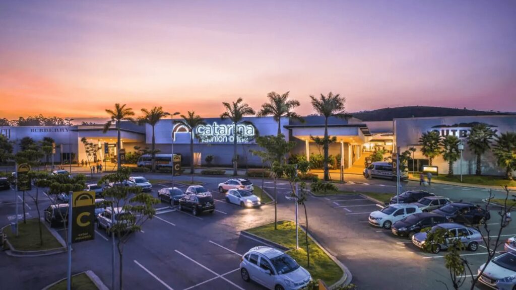 Crepúsculo sobre um shopping center com sinalização iluminada e estacionamento lotado.