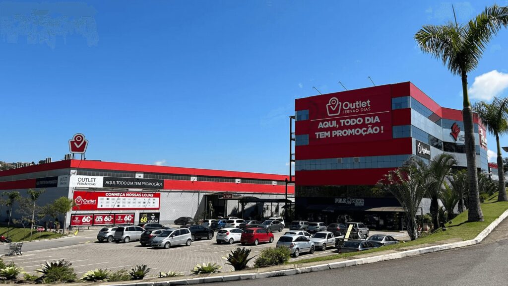 Edifício de shopping center vermelho e preto com carros estacionados e palmeiras sob um céu azul claro.