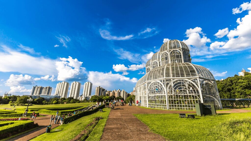 Estufa de vidro em um parque com céu azul, nuvens e horizonte da cidade ao fundo.