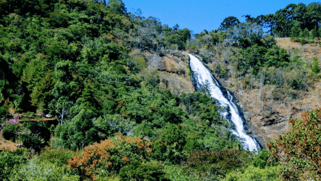 Uma cachoeira alta e imponente despenca por uma encosta verdejante em meio a uma floresta densa. Uma pequena construção pode ser vista ao longe, no canto inferior esquerdo da imagem.