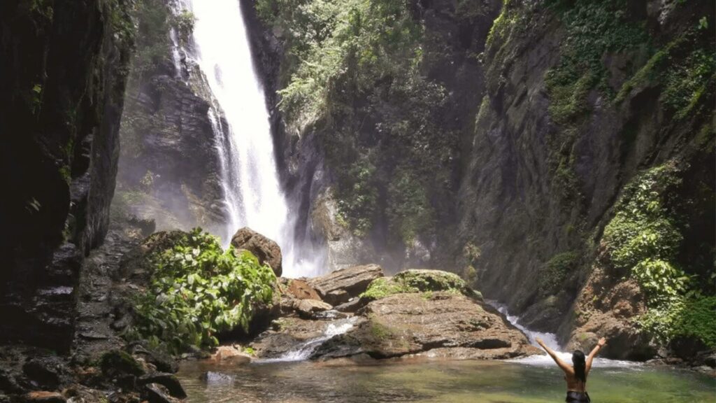 Uma cachoeira alta e imponente desce por uma encosta íngreme e rochosa, cercada por vegetação exuberante. A água cai em várias camadas, criando uma névoa refrescante e um som relaxante. A cachoeira está localizada em uma área natural preservada, com árvores verdes e flores coloridas.