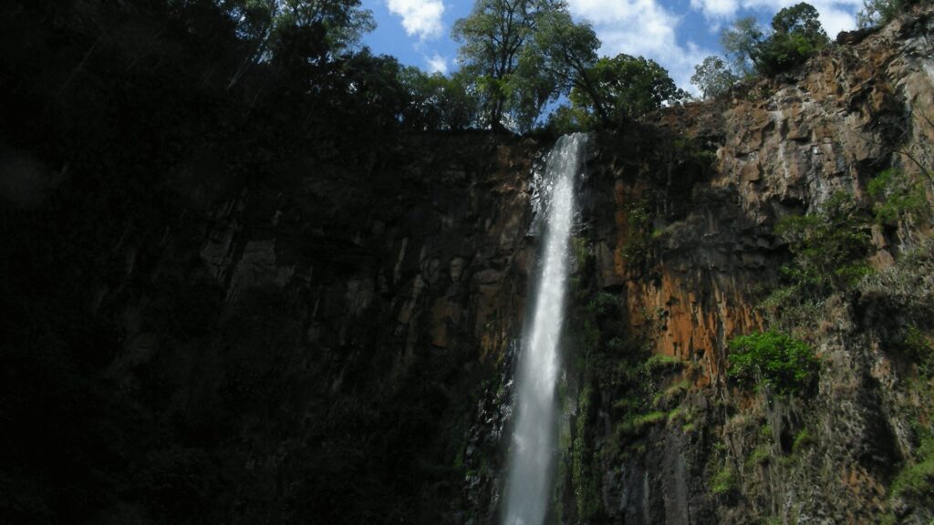 Uma cachoeira estreita e alta cai de um penhasco íngreme e rochoso, formando uma cortina de água branca e espumante. A água desce com força, contrastando com a rocha escura e úmida. A vegetação verdejante se agarra às laterais do penhasco, adicionando vida e cor à cena. O topo da cachoeira é emoldurado por um céu azul com nuvens brancas e algumas árvores. A imagem transmite a grandiosidade e a beleza da natureza intocada.