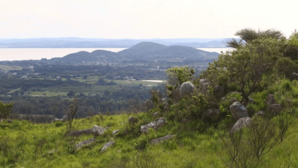 Colinas onduladas com vegetação e rochas espalhadas, com vista para um lago distante.